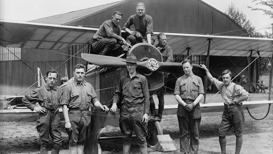A black and white photo of men in Army uniforms standing near and atop an early 20th-century propeller plane